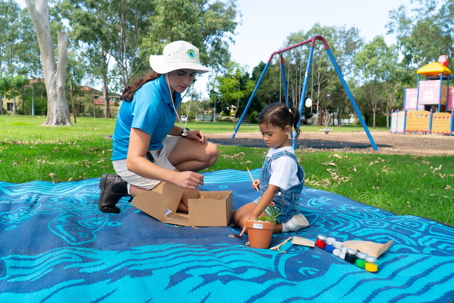 Sabrina Guse from EnviroMentoring is teaching a young girl how to complete her EnviroMentoring Paint and Plant DIY kit. She is excitedly pointing at a bug on the recycled blue mat.