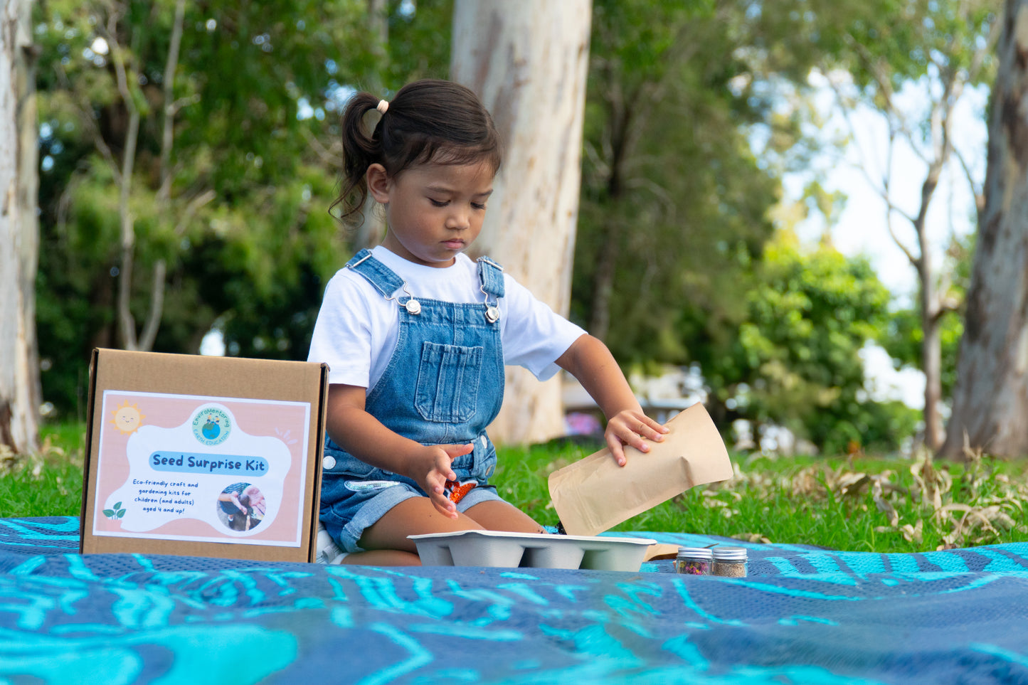 A young girl is pouring the materials into her tray to make her EnviroMentoring Seed Surprise DIY Kit. The EnviroMentoring Seed Surprise DIY Kit is in view and the girl is sitting on a mat outside.