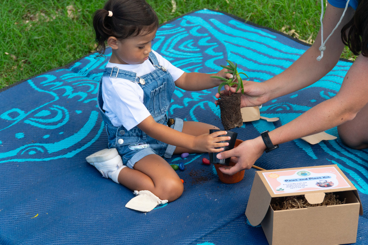 Young girl being shown by Sabrina Guse from EnviroMentoring how to repot her native pigface succulent into her terracotta pot from her EnviroMentoring Paint and Plant DIY kit.