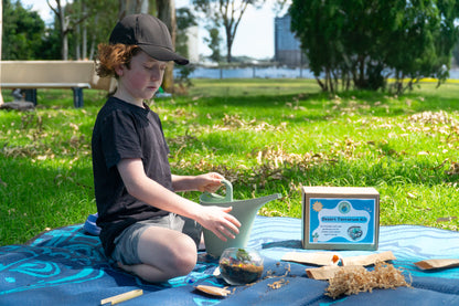 A young boy waters his completed EnviroMentoring Desert Terrarium DIY Kit. The box is in the background.