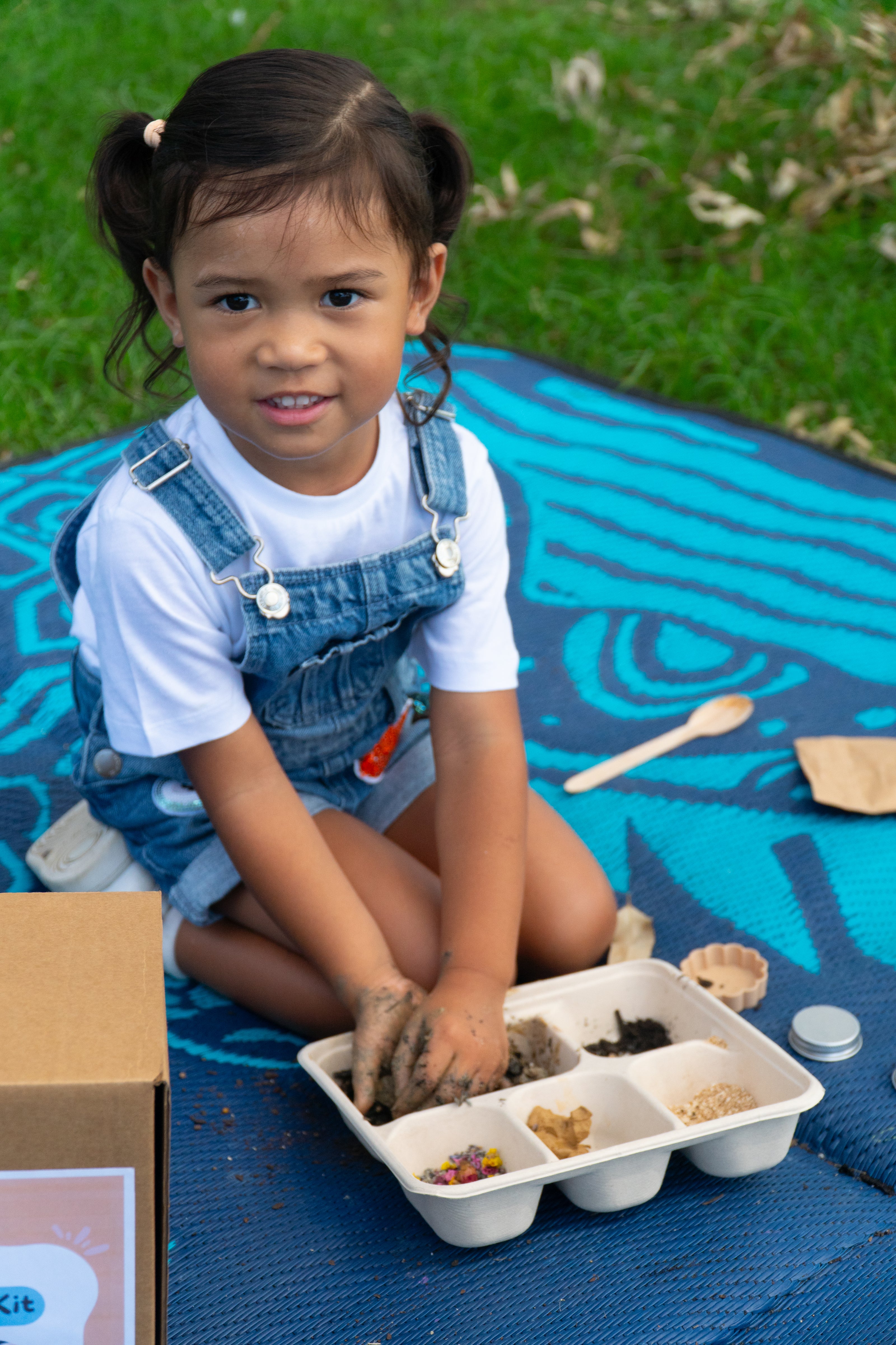 A girl is mixing soil, clay, flowers and seeds to make a seed surprise from an environmental Seed Surprise DIY Kit. She is sitting on a mat, smiling up at the camera.