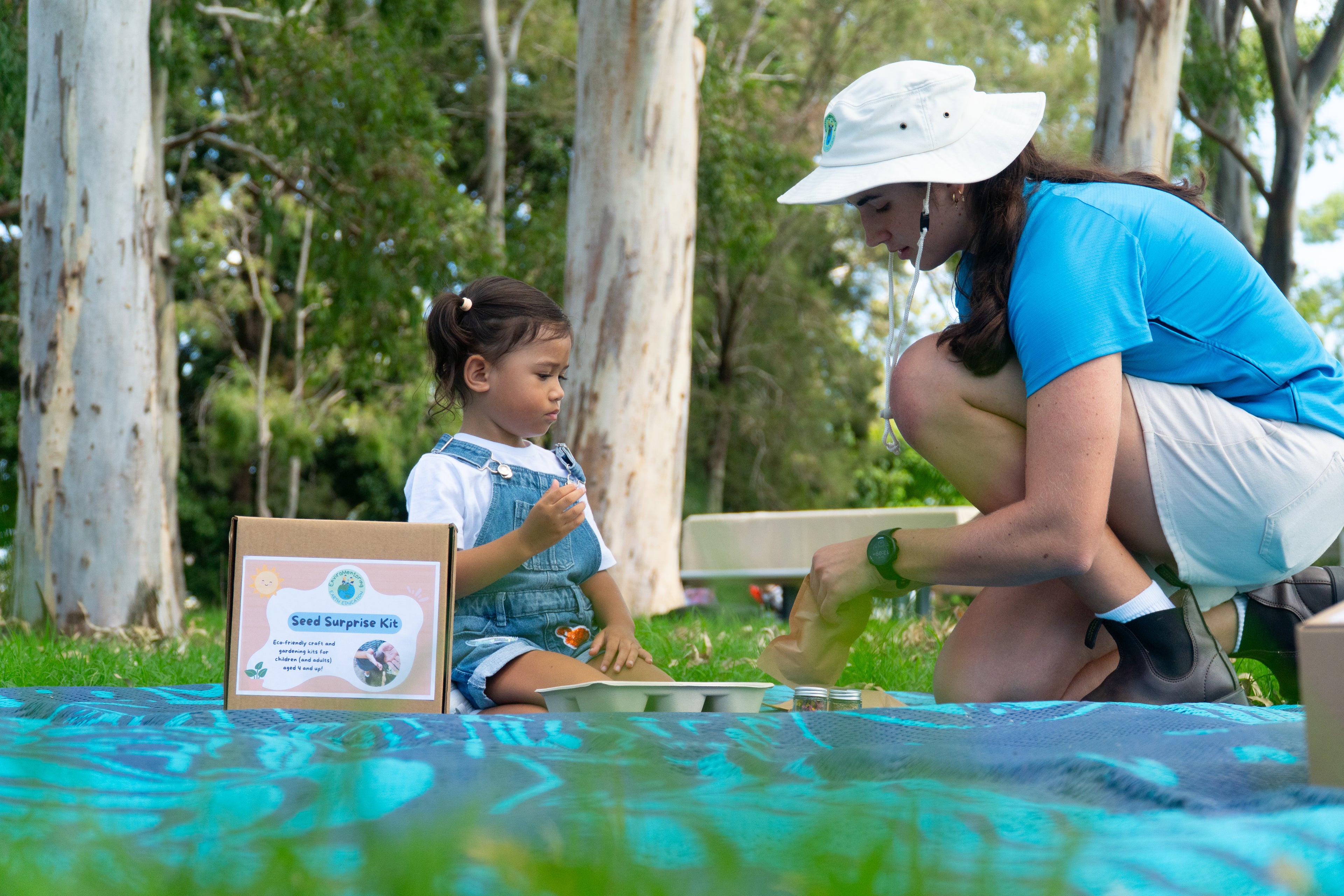 A young girl is being taught by Sabrina Guse from EnviroMentoring how to make her seed surprise. A box with the Seed Surprise DIY Kit is in view.
